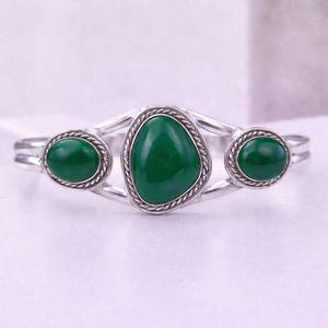 Op de afbeelding: Een zilverkleurige armband met drie heldergroene edelstenen. De centrale steen heeft een onregelmatige vorm, terwijl de zijstenen cirkelvormig zijn. Elke steen is omlijst door een gedraaide, touwachtige rand.