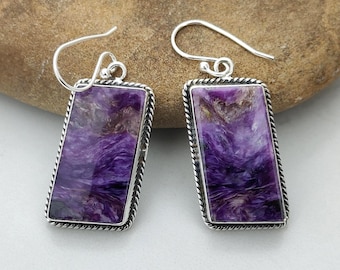 May include: A pair of silver earrings with rectangular, purple stone pendants. The stones have a swirling pattern of purple and white. The earrings have a decorative silver rope-like border and silver ear wires.