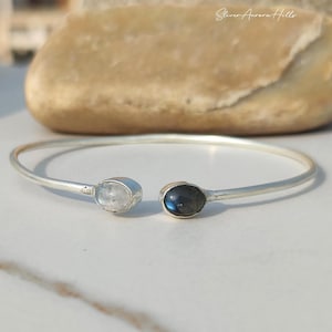 May include: A silver-coloured open cuff bracelet featuring a light blue and a dark blue oval gemstone at each end. The bracelet is displayed on a white surface with a large, light brown rock in the background.