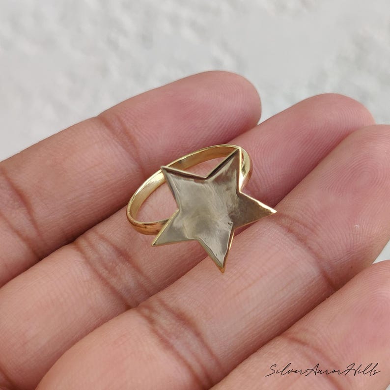May include: A gold-toned ring featuring a flat, five-pointed star centerpiece with a smooth, reflective surface. The star is attached to a thin, circular band, creating a simple and clean design that sits prominently on the finger.