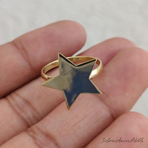 May include: A gold-toned ring featuring a prominent, flat, five-pointed star centerpiece. The star has a smooth, reflective surface and is attached to a simple, thin, circular band. The text Silver Aurora Hills is visible in the bottom right corner.
