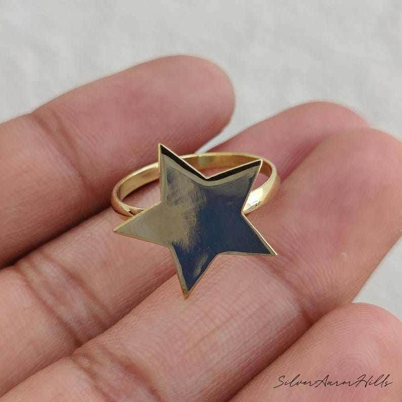May include: A gold-toned ring featuring a prominent, flat, five-pointed star centerpiece. The star has a smooth, reflective surface and is attached to a simple, thin, circular band. The text Silver Aurora Hills is visible in the bottom right corner.