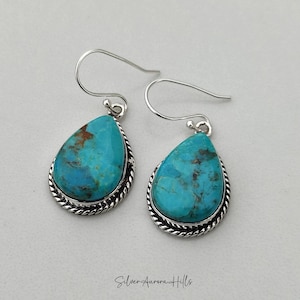 May include: A pair of turquoise teardrop earrings with a silver rope-like border and silver ear hooks. The turquoise stones have brown inclusions. The earrings are displayed on a light gray background. The text "Silver Aurora Hills" is at the bottom.