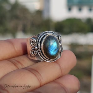 May include: A silver ring with an oval, iridescent labradorite stone. The ring has an ornate design with scrollwork and a wide band. The stone displays blue and green flashes. The ring is held up by a hand.