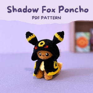 Może przedstawiać: Szydełkowa pluszowa figurka lisa w czarnym ponczo z żółtymi wykończeniami i pasującym kapturem. Kaptur ma żółte paski i żółte koło. Tekst "Shadow Fox Poncho PDF Pattern" znajduje się nad figurką.