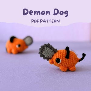 Puede incluir: Dos figuras de crochet naranja de un perro con cabeza de motosierra, detalles negros y ojos grandes negros. El texto "Demon Dog PDF Pattern" se muestra en púrpura sobre las figuras sobre un fondo lavanda.