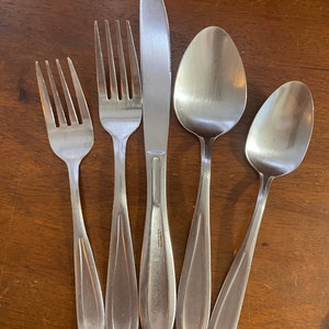 May include: A set of stainless steel silverware including two forks, a knife, and two spoons. The utensils are arranged on a wooden surface. The knife has a serrated edge. The spoons have oval bowls.
