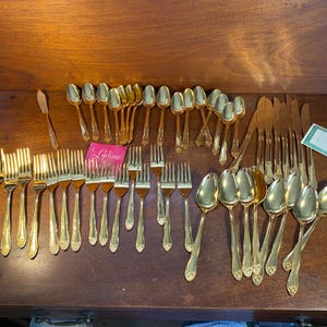 May include: A collection of gold-colored flatware, including forks, spoons, knives, and a butter knife, arranged on a wooden surface. The flatware has decorative handles and is likely part of a dining set. A pink card with the word "Lifetime" is visible.