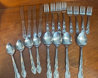 May include: A collection of stainless steel silverware, including knives, forks, and spoons, arranged on a wooden surface. The silverware has a decorative handle design.