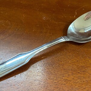 May include: A silver spoon with a rounded bowl and a rectangular handle. The handle has a raised design.
