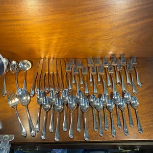 May include: A collection of polished silverware, including forks, spoons, knives, and serving spoons, arranged on a wooden surface. The silverware has a reflective, silver finish, and the arrangement suggests a formal dining setting.