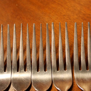 May include: Eight silver forks lined up in a row on a wooden surface. The forks are all the same size and shape, with three prongs each.
