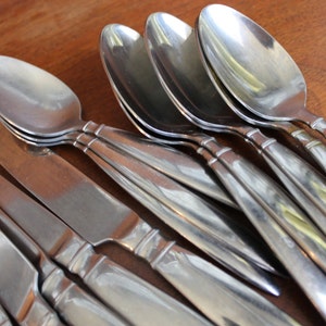 May include: A set of silver cutlery, including spoons and knives, arranged in a neat stack. The cutlery is shiny and reflects light.