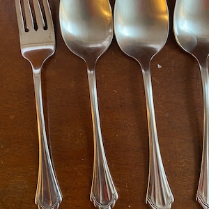 May include: A set of stainless steel silverware, including a fork and three spoons, arranged on a wooden surface. The silverware has a decorative, scalloped design at the base of the handles.