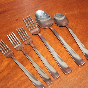 May include: A set of stainless steel silverware, including forks and spoons, arranged diagonally on a wooden surface. The forks have four tines, and the spoons have oval bowls. The silverware has a simple, modern design.