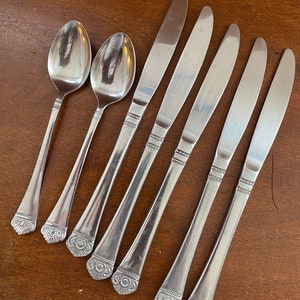 May include: Set of seven silver-toned flatware pieces, including two spoons and five knives. The handles of the flatware are decorated with a floral design.