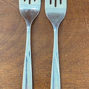 May include: Two stainless steel forks with four tines each, arranged vertically on a wooden surface. The handles have a subtle, curved design. The forks are clean and reflect the light.