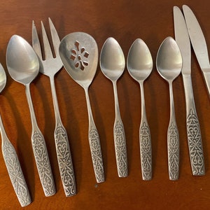 May include: A set of eight silver-toned flatware pieces with ornate designs. The set includes a serving spoon, a serving fork, a butter knife, and five spoons.
