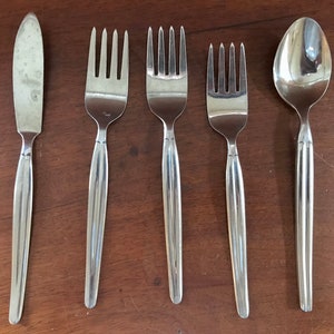 May include: A set of stainless steel silverware including a butter knife, three forks, and a spoon. The utensils have long, smooth handles and are arranged on a dark wooden surface. The silverware is clean and reflects the light.