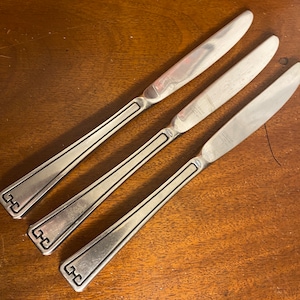 May include: Three silver dinner knives with a decorative pattern on the handle. The knives have a simple, elegant design.