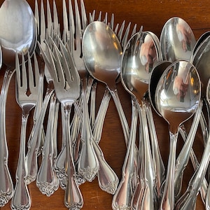 May include: A collection of shiny, silver-colored silverware, including forks and spoons, arranged on a wooden surface. The silverware has a decorative floral design on the handles. The spoons and forks are of various sizes.