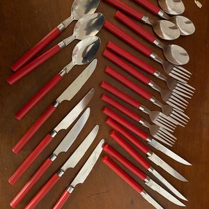 May include: A set of red handled silverware, including spoons, forks, and knives. The silverware is laid out on a wooden surface.