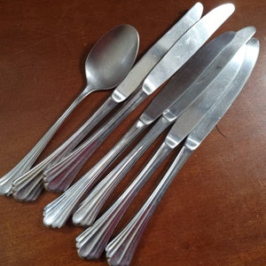 May include: A set of stainless steel silverware, including a spoon and several knives, arranged on a brown wooden surface. The silverware has a decorative, ridged handle design.