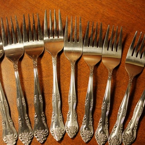 May include: A set of ten silver forks with ornate handles. The forks are arranged in a row on a wooden surface.
