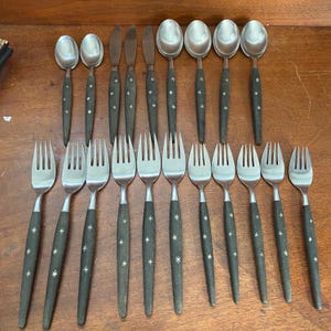 May include: A collection of stainless steel flatware featuring dark charcoal handles with a white starburst pattern. The set includes various spoons, forks, and knives arranged in rows, showcasing a consistent design with metallic heads and dark handles.