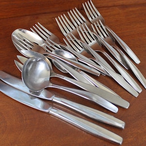 May include: A set of silverware, including forks, spoons, and a ladle, all made of polished stainless steel. The silverware is arranged in a pile on a wooden surface.