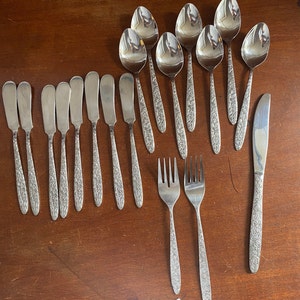 May include: A set of 14 silver-toned utensils, including spoons, forks, and knives, with ornate floral designs. The utensils are arranged on a wooden surface.
