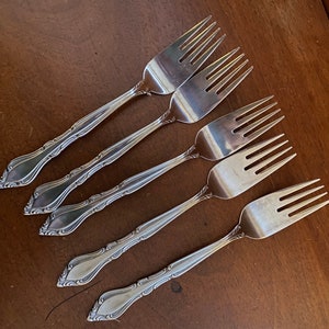 May include: Five silver forks with ornate handles. The forks have a decorative design on the handle and are arranged in a row.