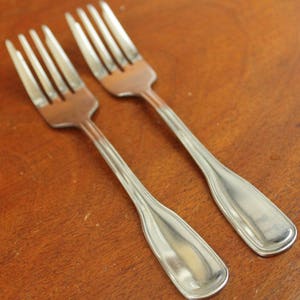 May include: Two silver forks with a polished finish. The forks are laid out side by side on a wooden surface.