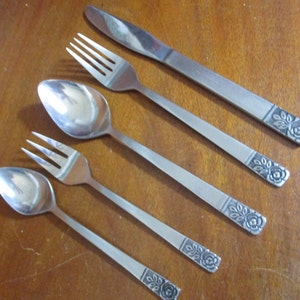 May include: Set of four silver-toned flatware pieces with rose-shaped designs on the handles. The set includes a knife, two forks, and a spoon.