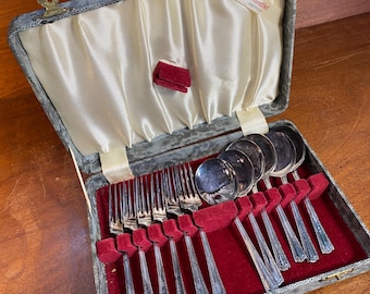 Op de afbeelding: Een vintage zilveren bestekset in een grijs fluwelen etui met een rode fluwelen binnenkant. De koffer heeft een messing sluiting en een label met de tekst "De Montfort Sheffield".