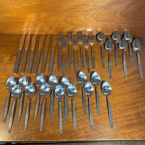 May include: A set of stainless steel silverware, including forks, knives, and spoons. The silverware is arranged in rows on a wooden surface.