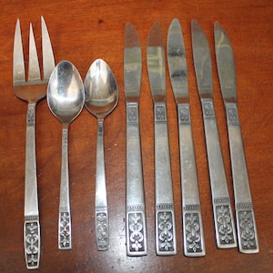 May include: A set of eight silver-toned flatware pieces, including a fork, two spoons, and five butter knives. Each piece features a decorative floral design on the handle.