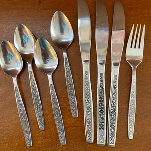 May include: A set of eight silver-toned flatware pieces with floral designs. The set includes four spoons, three knives, and one fork.