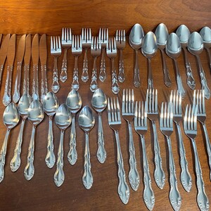 May include: A collection of stainless steel silverware, including knives, forks, and spoons, arranged on a wooden surface. The silverware has a decorative floral pattern on the handles.