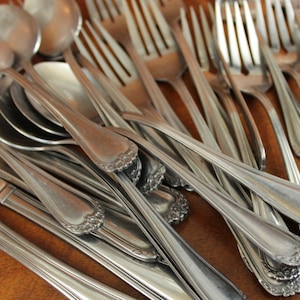 May include: A pile of silver cutlery, including forks and spoons, with ornate handles. The cutlery is arranged in a random pattern, with some pieces overlapping.