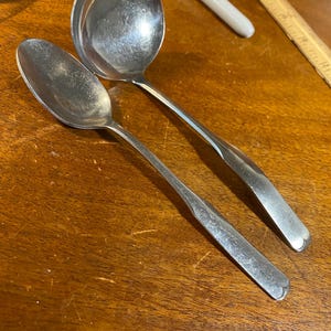 May include: Two silver serving spoons, one a standard spoon and the other a ladle, are lying on a wooden surface.