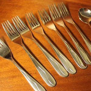 May include: A set of eight silver-toned forks and a spoon with a wavy design on the handles. The forks and spoon are arranged in a row on a wooden surface.
