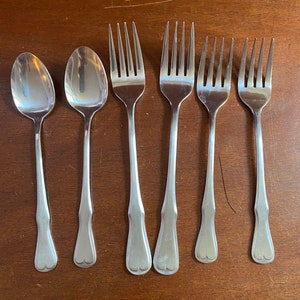 May include: A set of six stainless steel silverware pieces, including two spoons and four forks. The silverware has a simple, elegant design with a heart-shaped detail at the base of the handles. The pieces are arranged on a dark brown wooden surface.