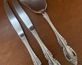May include: Three silver-toned, ornate, decorative silverware pieces: two butter knives and a sugar spoon. The knives have a floral design on the handles. The spoon has a scalloped edge and a floral design on the handle.