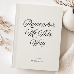 May include: An off-white hardcover book with the title "Remember Me This Way" in a decorative script. The author's name, Laurel Anne, is printed below the title. The book is on a white surface, with a small vase of dried flowers in the background.