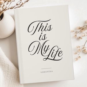 May include: A cream-colored journal with the words "This is My Life" in elegant black script. The name "Samantha" is printed below the title. The journal is placed on a white surface with decorative dried flowers.