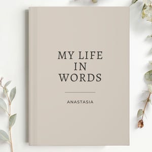 May include: A beige hardback journal with the text "MY LIFE IN WORDS" and the name "ANASTASIA" printed in black. The journal is surrounded by sprigs of greenery.