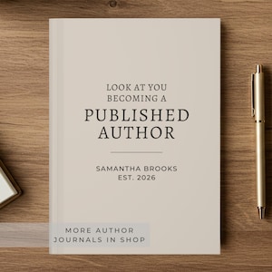 Peut inclure: Un journal beige avec le texte "LOOK AT YOU BECOMING A PUBLISHED AUTHOR" en noir. En dessous, on peut lire "SAMANTHA BROOKS EST. 2026". Un stylo doré repose sur une surface en bois à côté du journal. Le journal est un livre.