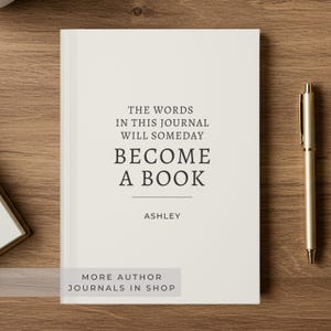 May include: A cream-colored journal with the text "THE WORDS IN THIS JOURNAL WILL SOMEDAY BECOME A BOOK" and the name "ASHLEY" printed on the cover. A gold pen rests beside the journal on a wooden surface. The journal is a hardcover book.