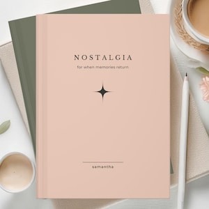 Op de afbeelding: Een roze notitieboek met het woord "NOSTALGIA" en de tekst "for when memories return" op de kaft. Een zwarte sterrenburst-afbeelding is gecentreerd onder de tekst. De naam "samantha" staat onderaan gedrukt. Een witte pen ligt ernaast.